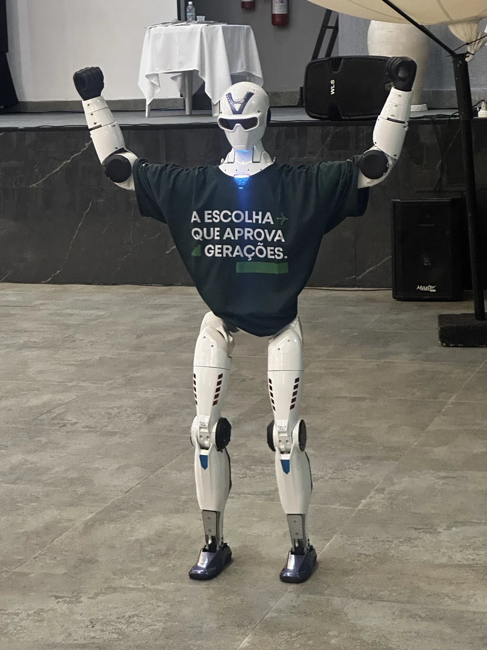 Robô humanoide R1 com os braços erguidos, vestindo a camiseta da Convenção Interna do Colégio COC Paulínia, um evento interno de empresa